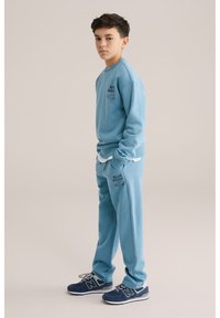 Svetlo modra majica s kapuco in ustrezne trenirke z logotipom, v kombinaciji z mornarskimi športnimi copati. Mehka tkanina z zasnovo sproščenega kroja.