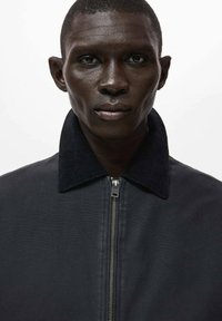 Čierna bunda s golierom z manžestru, s predným zipsom a hladkou textúrou látky. Jednoduchý dizajn bez viditeľných vzorov alebo doplnkov.