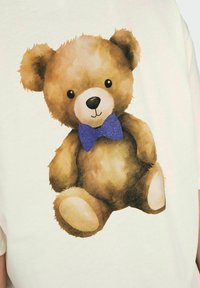 T-shirt imprimé mettant en avant un ours en peluche brun en cartoon avec un nœud papillon bleu, texture douce, fond clair, et yeux ronds et brillants noirs.