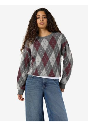 Vrouw met lang krullend haar, gekleed in een grijze en bordeauxrode argyle-trui en blauwe jeans, staand tegen een effen lichte achtergrond.