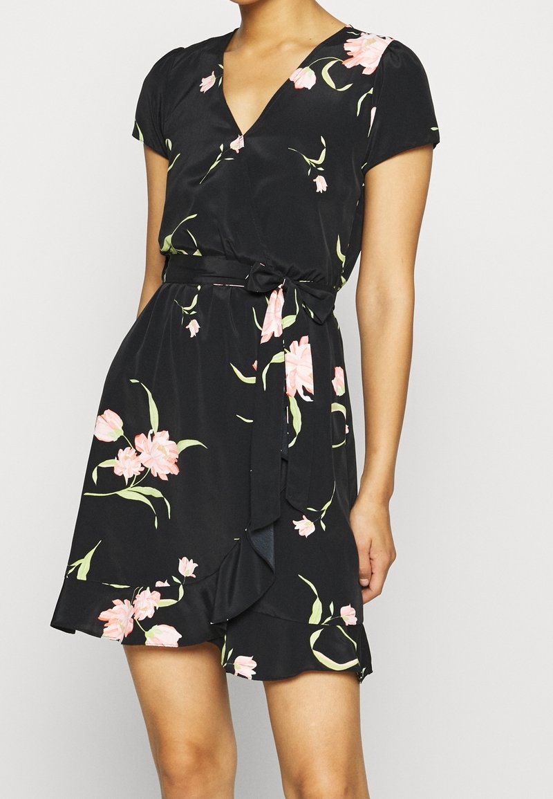Femme portant une courte robe portefeuille noire avec des motifs floraux roses et un nœud noué à la taille, debout devant un fond clair uni.