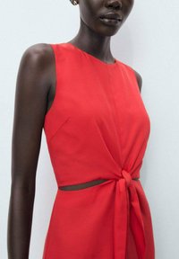 Red sleeveless dress with a fitted bodice, tie-front detail, and cut-out at the waist. Made of smooth fabric with a soft texture.