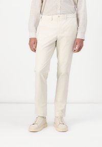 Pantalones de algodón beige con un corte recto, que cuentan con trabas para el cinturón y un cierre de botón. Combinados con zapatillas de color claro.