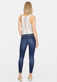 Jeans oscuros ajustados y delgados con dos bolsillos traseros, combinados con una camiseta sin mangas blanca y texturizada. El modelo está de espaldas mirando hacia la cámara.