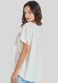 Blusa blanca de manga corta con un corte holgado, tejido suave, escote redondo y sutil detalle de encaje en los hombros. Vista posterior mostrada.