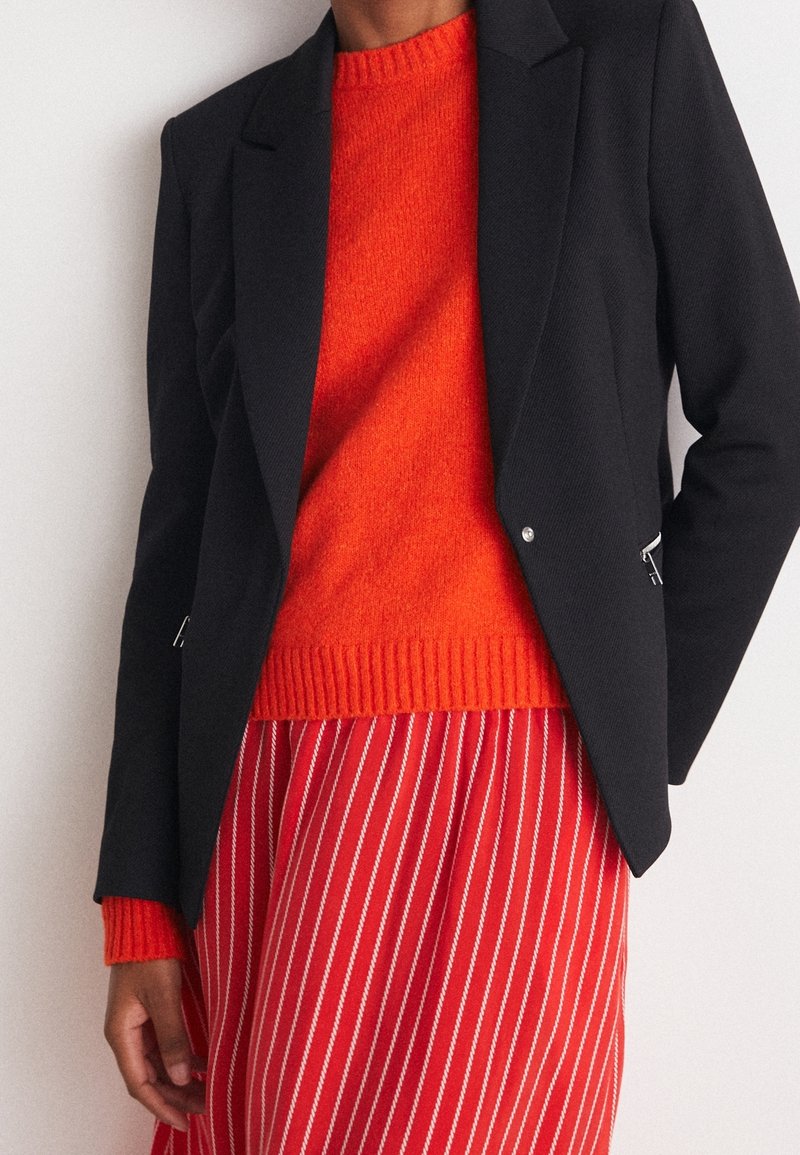 Blazer nero con rever affilati, abbinato a un maglione rosso a maglia e una gonna a righe. Il blazer presenta dettagli in metallo argentato.