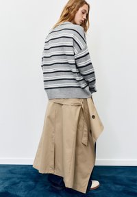 Gestreepte grijs-witte trui gecombineerd met een beige trenchcoat. De jas heeft knopen, een ceintuur en een vloeiende pasvorm.