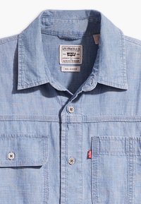 Camicia in denim azzurro chiaro con vestibilità rilassata, colletto con bottoni, due tasche frontali e dettagli metallici con marchio, completi di un'etichetta rossa.