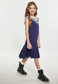 Marineblaues Kleid mit V-Ausschnitt und weißen Streifenakzenten, mit einem ausgestellten Rock und strukturiertem Stoff. Kombiniert mit schwarzen Schnürstiefeln.