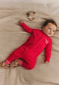 Rød ribbet Onepiece med en frontlynlås og "santa baby" tryk, parret med stribede manchetter. En blød hundelegetøj ligger tæt på på et struktureret tæppe.