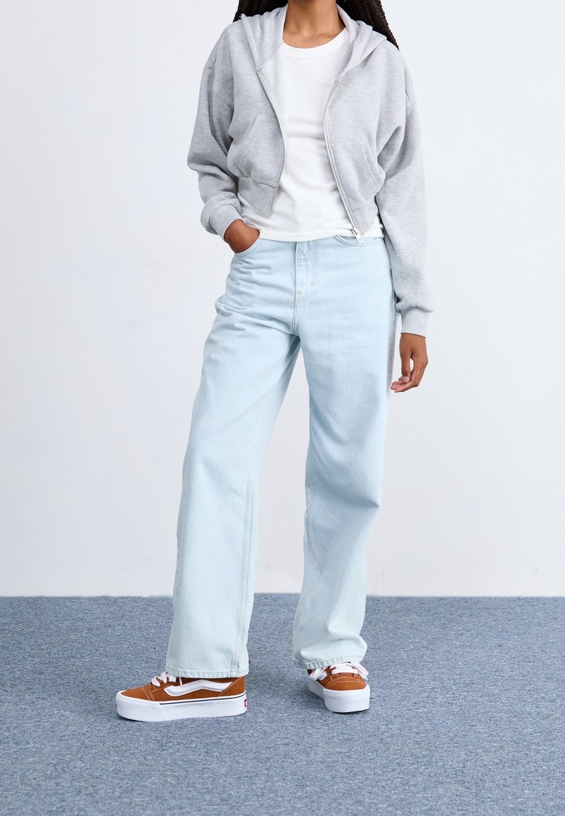 Personne portant un sweat à capuche gris clair zippé, une chemise blanche, un jean large bleu clair et des baskets marron et blanches, debout sur un tapis bleu.