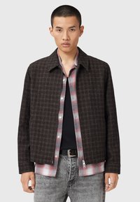 Veste à carreaux bruns avec col, fermeture zippée et poches latérales, superposée à une chemise à carreaux rouges, associée à un jean gris.