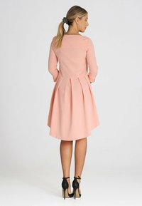 Helles rosa Kleid mit langen Ärmeln, plissiertem Rock und quadratischem Rückenausschnitt. Getragen mit schwarzen High-Heels. Glatte Stoffstruktur.