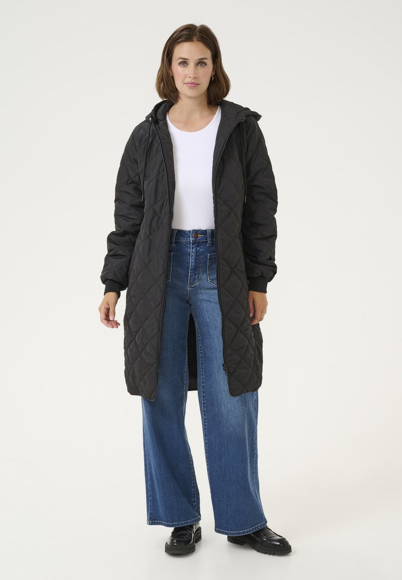 Manteau long noir matelassé avec capuche, doté d'une fermeture éclair sur toute la longueur, de poignets côtelés et d'un design jusqu'aux genoux, associé à un jean en denim bleu à jambes larges.