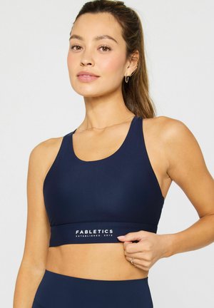 Frau trägt einen marineblauen Sport-BH mit dem Logo "Fabletics", steht vor einem schlichten hellen Hintergrund und hält den unteren Saum des BHs.