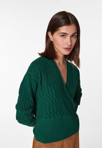 Grüner gestrickter Wickelsweater mit einem Zopfmuster, tiefem V-Ausschnitt und geripptem Saum sowie Bündchen, hergestellt aus einem weichen, strukturierten Stoff.