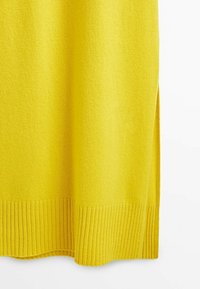 Keltainen neulepusero, jossa on ribbaushelma ja sivuhalkiot, pehmeällä tekstuurilla ja mukavalla laskoksella.