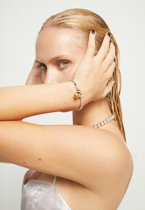 Femme aux cheveux blonds mouillés tenant sa tête avec ses deux mains, portant un bracelet en argent avec un charme en or et un collier de perles.