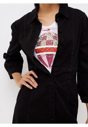 Mujer con chaqueta negra abotonada sobre una camiseta blanca con un diseño de globo aerostático rosa y rojo con lentejuelas.