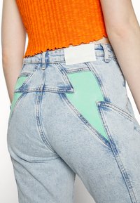 Blue jeans de denim em azul claro com um acabamento texturizado, apresentando detalhes verdes e um design triangular nos bolsos traseiros. Etiqueta branca na cintura.