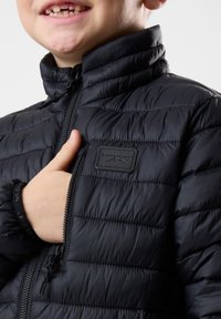 Sort isoleret jakke med quiltet design, høj krave og lynlås foran. Imponeret med et mærke på brystet og elastiske manchetter.