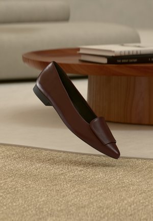 Scarpa bassa in pelle marrone scuro senza lacci che fluttua sopra un tappeto beige testurizzato, con un tavolino da caffè in legno e libri impilati sullo sfondo.