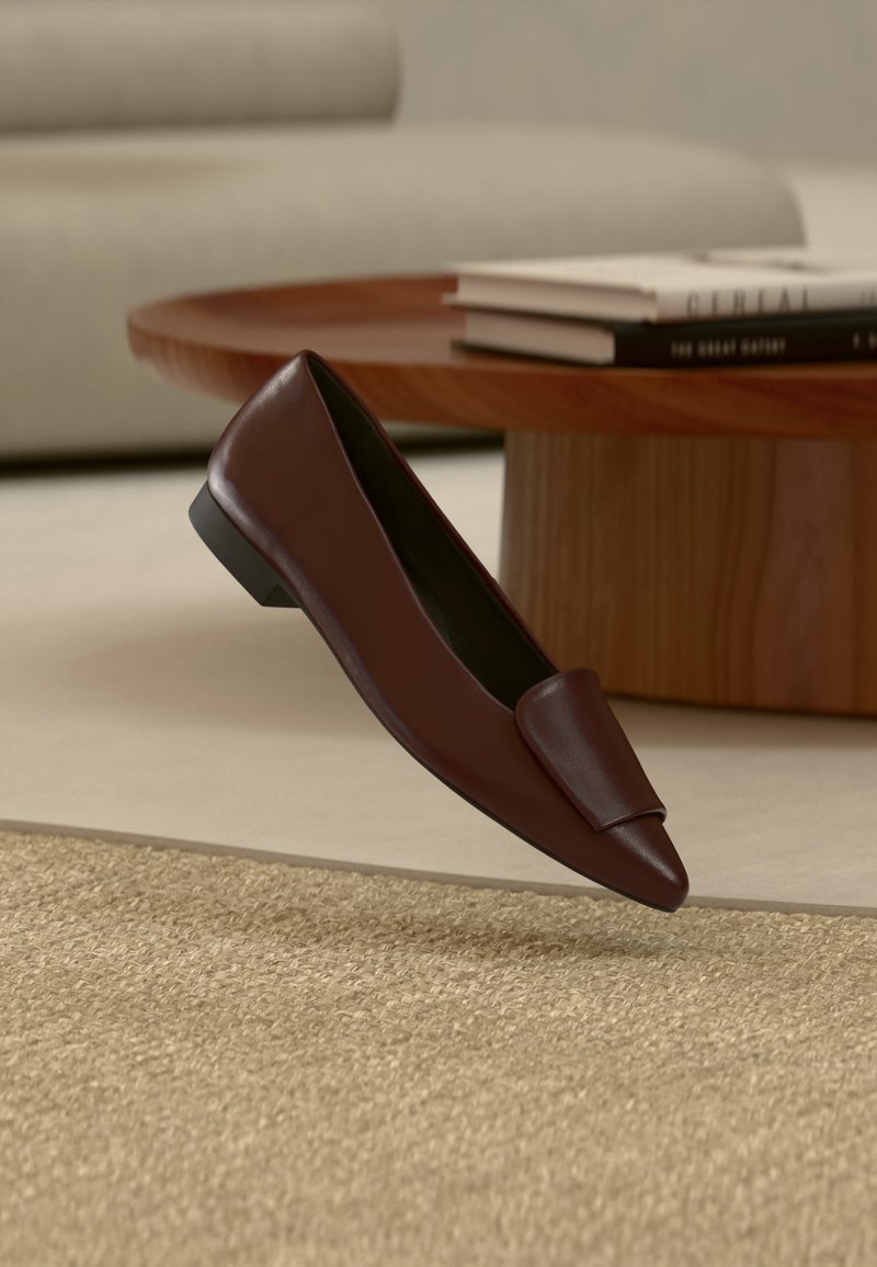 Σκούρο καφέ δερμάτινο παπούτσι τύπου slip-on με επίπεδη σόλα να αιωρείται πάνω από μπεζ υφασμάτινο χαλί, με ένα ξύλινο τραπεζάκι καφέ και στοιβαγμένα βιβλία στο φόντο.