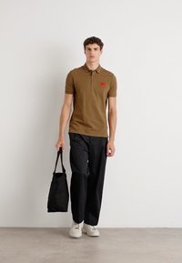Cămașă polo maro cu o patch-uri roșii, asortată cu pantaloni negri și adidași albi, ținând în mână o geantă tote neagră.