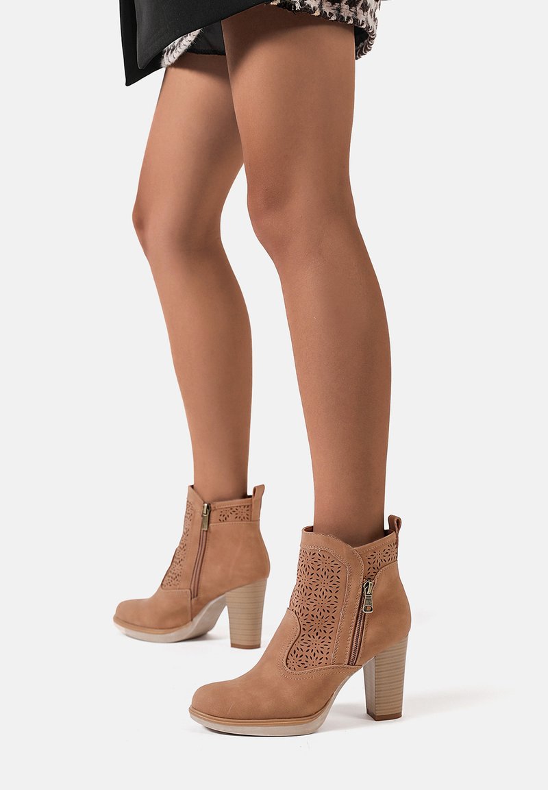 Stivaletti alla caviglia beige in suede, con un motivo decorativo intagliato, cerniere laterali e tacco a blocco. Texture liscia e punta arrotondata.