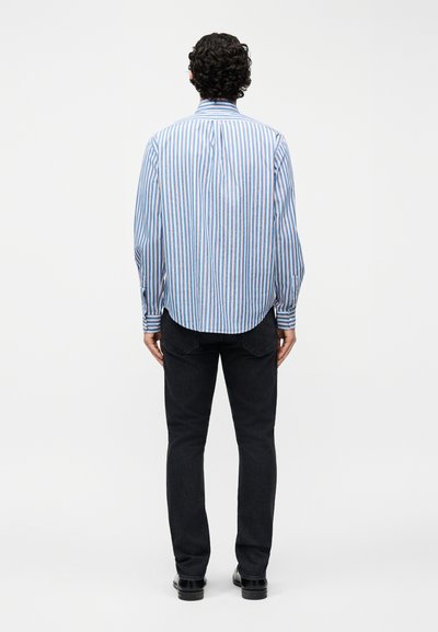 Άνδρας με σγουρά μαλλιά στέκεται με την πλάτη γυρισμένη, φοράει γαλάζιο και λευκό ριγέ πουκάμισο με κουμπιά, μαύρο τζιν και μαύρα παπούτσια μπροστά σε λευκό φόντο.