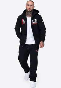 Sudadera negra con cremallera y logotipo rojo y blanco, combinada con pantalones de chándal negros con un pequeño logotipo, llevada con zapatillas blancas. Ropa deportiva casual.