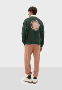 Felpa verde con grafica a raggi di sole rosa, abbinata a pantaloni della tuta rosa chiaro. Presenta polsini elastici e sneakers bianche con suole marroni.