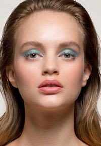 Porträtt av en person med slät hy, iförd turkos ögonskugga och ljusrosa läppfärg, med långt, något vågigt hår.