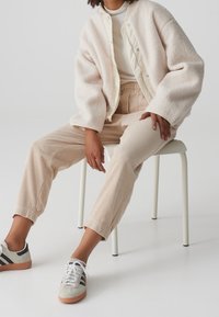 Personne assise sur un tabouret blanc portant une veste sherpa crème, un pantalon en velours côtelé beige, des baskets blanches avec des bandes noires, et un haut blanc.