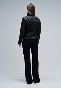 Frau steht mit dem Rücken zur Kamera, trägt eine schwarze gesteppte Jacke mit Chevron-Steppung und schwarze weit geschnittene Hosen vor einem schlichten grauen Hintergrund.