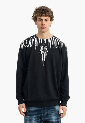 Jeune homme aux cheveux bouclés portant un sweat-shirt noir avec un texte blanc déchiqueté et un motif d'éclair sur les épaules et la poitrine.