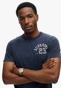 Dunkelblaues Rundhals-T-Shirt mit gesticktem "SUPERDRY 23" in Grau, weiche Textur, getragen mit einer braunen Lederuhr am Handgelenk.