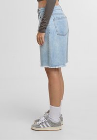 Persona con falda de mezclilla azul clara, camiseta de manga larga gris, calcetas blancas y zapatillas Adidas grises, de pie sobre un fondo blanco liso.