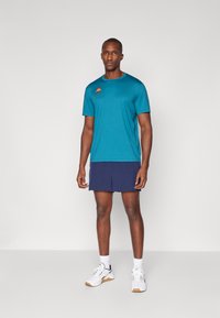 Mannen sportoutfit met een teal T-shirt met korte mouwen en een logo, gecombineerd met marineblauwe shorts. Draagt met witte sneakers en enkelsokken.