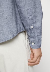 Texturovaná modrá košile s dlouhými rukávy, knoflíkovými manžetami a zakřiveným lemem, kombinovaná s světlými manšestrovými kalhotami.