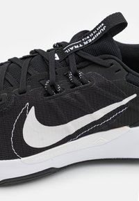Zapatilla deportiva negra con parte superior de tela, Swoosh blanco de Nike, acentos texturizados y cordones negros; incluye una lengüeta en el talón y una media suela blanca con marca.