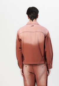 Rust-colored denim jacket with a faded gradient, featuring embroidery on the back and button closure at the cuffs.