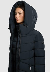 Marineblaue Steppjacke mit großer Kapuze, ausgestattet mit einem Reißverschluss, genieteter Textur und einem logobeschrifteten Akzent am Ärmel. Leicht und isoliert.