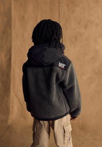 Person with dreadlocks wearing a gray and black Superdry fleece jacket and beige cargo pants, facing away against a brown backdrop.