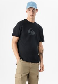 Ung mann står selvsikkert iført en svart T-skjorte med logo, khaki cargo-bukser og en lys blå caps, mot en ensfarget hvit bakgrunn.