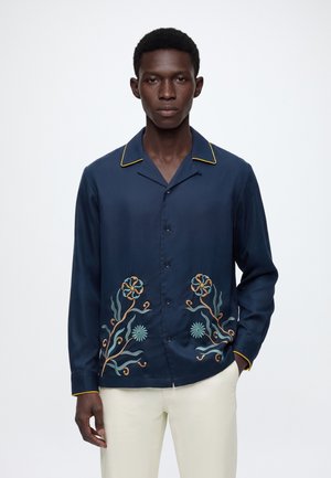 Homme portant une chemise bleu foncé à boutons avec une bordure jaune et une broderie florale sur le devant, associée à un pantalon beige clair.