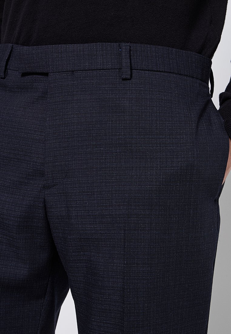 Los pantalones de color azul marino texturizados presentan un ajuste entallado, frente plano y presillas para cinturón, exhibiendo sutiles rayas verticales en la tela.
