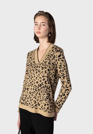 Jeune femme debout portant un pull beige à col en V imprimé léopard et un pantalon noir sur un fond gris clair uni.