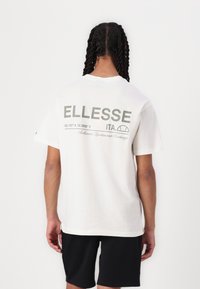 Biele bavlnené tričko s veľkým zeleným logom "ELLESSE" na chrbte, s doplňujúcim textom a súradnicami, krátkymi rukávmi a voľným strihom.