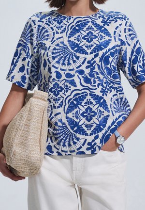 Femme portant un pantalon blanc et une chemise blanche à manches courtes avec des motifs floraux et d'éventail bleus complexes, tenant un sac beige tissé.